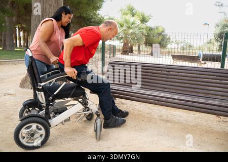 Une femme aide un homme à utiliser un fauteuil roulant lorsqu’il s’approche d’un banc dans un parc, soulignant l’inclusivité et l’accessibilité à l’extérieur, favorisant la communauté Banque D'Images