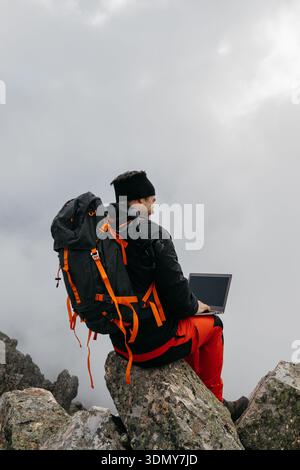 Un randonneur s'assoit sur un sommet rocheux, en utilisant un ordinateur portable sur fond de nuages et de ciel. La scène combine l'aventure avec la technologie dans un inspirin Banque D'Images