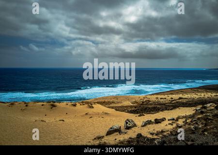 Une vaste étendue de l'océan bleu profond rencontre une côte sablonneuse sous un ciel spectaculaire et nuageux, avec des vagues qui s'écrasent à terre Banque D'Images