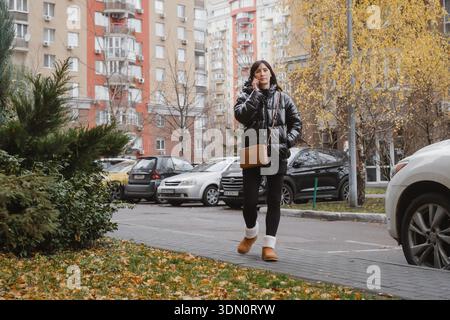 Femme souriante dans ses années 40 se promène dans la rue de la ville dans la veste gonflante brillante, parlant sur smartphone pendant la journée d'automne décontractée. Femelle aime amical Banque D'Images