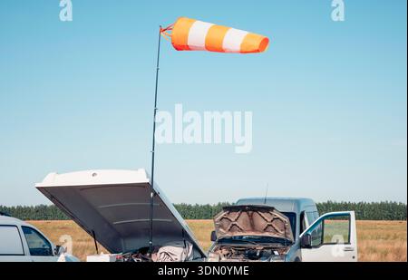 Une scène de ferme avec deux véhicules, un avec un capot ouvert. Une chaussette à vent est visible, indiquant la direction du vent. Le cadre est clair et ensoleillé, typique pour Banque D'Images