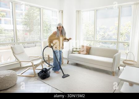 Femme avec de longs cheveux gris et des vêtements décontractés aspirant le tapis Banque D'Images