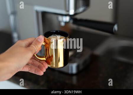Main de femme ramassant une tasse d'or de café fraîchement infusé à partir d'une machine à café maison argentée - routine matinale élégante, scène de cuisine confortable Banque D'Images