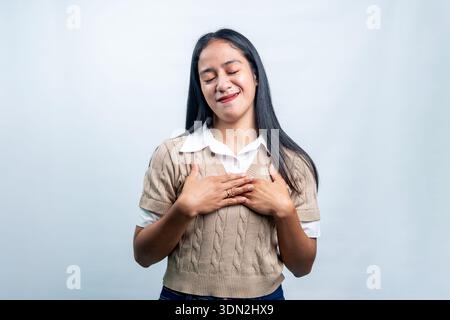 Une jeune femme asiatique les yeux fermés et les mains sur la poitrine, exprimant gratitude, paix et sincérité, isolée sur un fond blanc propre. Banque D'Images
