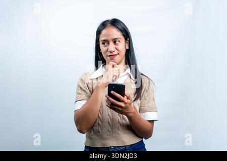 Une jeune femme asiatique regardant réfléchie tout en tenant un téléphone portable avec la main sur le menton. Isolé sur un fond lumineux pour la technologie et le shopping. Banque D'Images