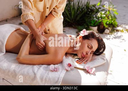 Femme recevant un massage professionnel du dos sur la table de spa à l'extérieur avec des fleurs d'orchidée et la lumière du soleil. Idéal pour la publicité bien-être, promotion Resort, an Banque D'Images