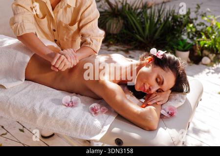 Femme recevant un massage professionnel du dos sur la table de spa à l'extérieur avec des fleurs d'orchidée et la lumière du soleil. Idéal pour la publicité de bien-être, le marketing de station, un Banque D'Images