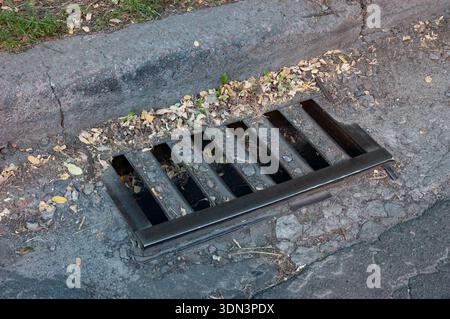 Vieille grille de drain d'orage en métal près du trottoir avec une petite feuille d'automne tombée à proximité. Détail urbain avec toucher saisonnier et texture de surface altérée. Banque D'Images