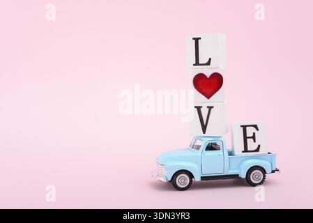 Une voiture jouet rétro bleue conduisant quatre cubes en bois épelant le mot Love sur un fond rose clair. Concept de la Saint-Valentin Banque D'Images