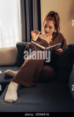 Une femme réfléchie est assise confortablement sur un canapé à la maison, tenant un cahier coloré et un stylo tout en réfléchissant pendant un moment de calme. La scène représente Banque D'Images