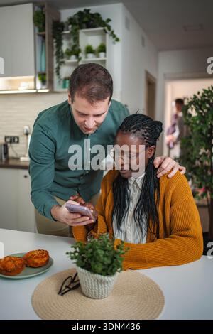 Couple diversifié à l'aide d'un smartphone à la table de cuisine à la maison Banque D'Images