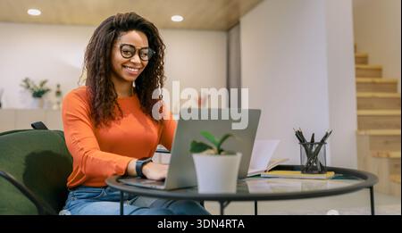 Jeune femme travaille sur ordinateur portable à table dans la salle moderne tout en souriant et en regardant concentré Banque D'Images