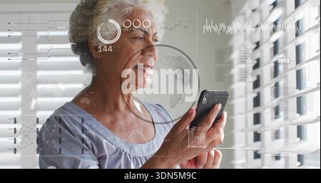 Tenant smartphone, femme senior en blouse bleu clair tapant écran à la maison par des stores, visionnant HUD Banque D'Images