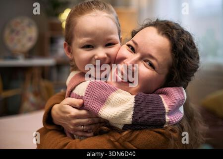 Femme caucasienne embrassant une fille caucasienne souriante à l'intérieur, les deux regardant la caméra, montrant un lien familial étroit et une expression joyeuse, visages proches les uns des autres Banque D'Images