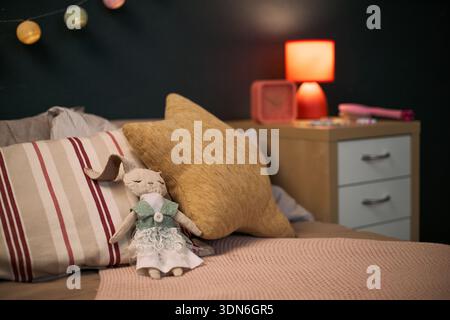Jouet de lapin en peluche portant robe reposant sur le lit avec des oreillers rayés et solides dans le cadre confortable de la chambre à coucher, table de nuit avec lampe et objets décoratifs visibles en arrière-plan Banque D'Images