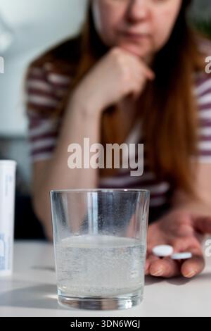 Une personne tenant deux pilules blanches est assise à une table, se préparant à prendre des médicaments. Un comprimé effervescent se dissout dans un verre d’eau, créant des bulles. Banque D'Images