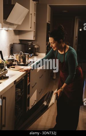 Femme dans un tablier charge un lave-vaisselle dans une cuisine moderne. Elle se concentre sur l'organisation de plats, créant une scène de la vie quotidienne à la maison. Cuisine applian Banque D'Images