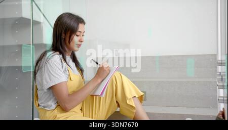 Écrivant femme asiatique dans une salopette jaune assise sur un escalier moderne, tenant un cahier en spirale et un stylo Banque D'Images