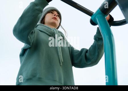 Prise de vue dynamique à faible angle regardant une jeune femme qui s'exerce sur une station de gym extérieure. La perspective souligne la structure de l'équipement, sa puissante pose et le ciel hivernal en arrière-plan. Banque D'Images