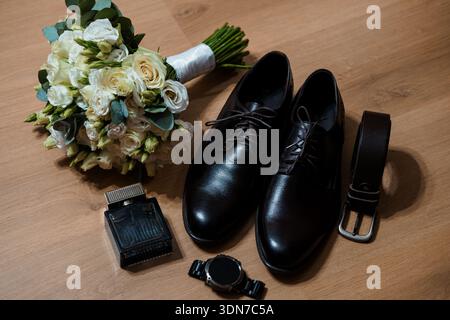 Un bouquet de mariage magnifiquement arrangé avec des roses blanches et de la verdure se trouve à côté de chaussures de robe noires, ceinture, montre, et bouteille de parfum sur un surfac en bois Banque D'Images
