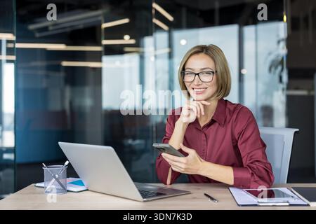 Femme d'affaires confiante souriant à la caméra, assise à son bureau moderne avec un ordinateur portable et tenant un smartphone, représentant le succès, la communication, la productivité et l'entreprise professionnelle Banque D'Images