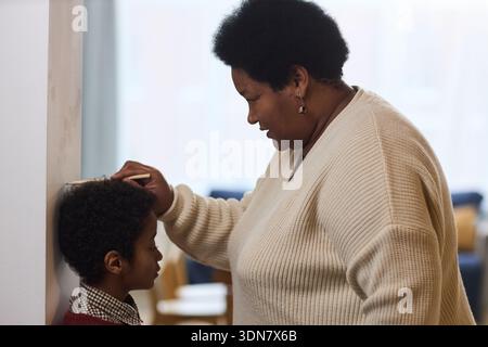 Femme noire d'âge moyen mesurant la hauteur d'un garçon d'enfant noir debout contre le mur, en utilisant la main et le crayon pour le marquage, les deux axés sur le processus, cadre familial intérieur Banque D'Images