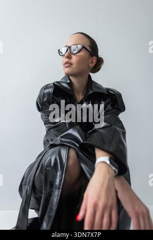 Portrait haute couture d'une jeune femme dans un trench-coat en cuir noir brillant et des lunettes de soleil avant-gardistes. Fond blanc minimaliste, chignon de cheveux élégant, Banque D'Images