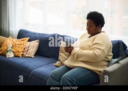Femme noire d'âge moyen assise sur un canapé à l'aide d'un smartphone, regardant l'appareil avec une expression focalisée, la lumière naturelle venant à travers la fenêtre en arrière-plan, cadre intérieur décontracté Banque D'Images