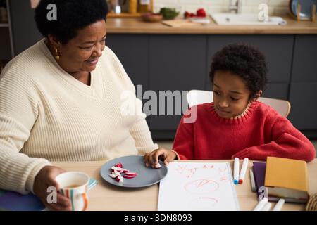 Femme senior noire assise avec un enfant noir dessinant sur papier à table, femme tenant une tasse et souriant tandis que l'enfant se concentre sur les œuvres d'art, les marqueurs de couleur et les livres sur la table Banque D'Images