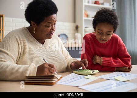 Femme noire d'âge moyen enseignant l'enfant à l'aide de la calculatrice et du cahier à table, les deux axés sur les documents financiers, l'enfant observant de près pendant que la femme écrit et calcule Banque D'Images