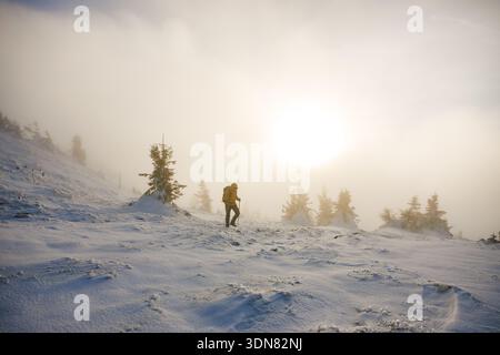 Randonnée en hiver. Randonneur marchant sur la crête de montagne enneigée pendant le coucher de soleil brumeux. Homme trekking en terrain extrême et par temps froid Banque D'Images