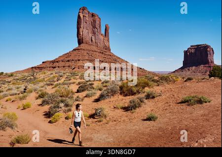 Une femme fait une randonnée à travers un paysage désertique avec des formations rocheuses massives sous un ciel bleu vif. Monument Valley, Arizona, États-Unis Banque D'Images