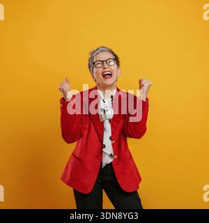 Happy Woman aux cheveux gris courts, vêtue d'un élégant blazer rouge et de lunettes, célèbre le succès avec les poings relevés contre un dos jaune vif Banque D'Images