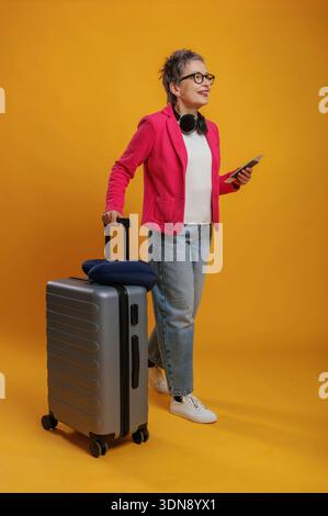 Femme âgée joyeuse portant un blazer rose et des lunettes élégantes tire une valise tout en tenant une tablette, exsudant l'excitation pour son prochain voyage Banque D'Images