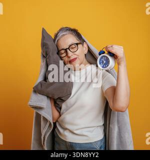 Femme aux cheveux gris portant des lunettes tient un oreiller et un réveil, exprimant la détente et le confort dans une atmosphère chaleureuse avec un jaune vif Banque D'Images