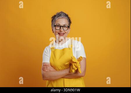 Femme âgée avec des cheveux gris et des lunettes, habillée d'un tablier jaune vif et de gants, se tient confiante avec les bras croisés contre un b jaune joyeux Banque D'Images