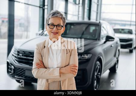 Femme mature aux cheveux gris courts, habillée d'un costume sur mesure, se tient en toute confiance dans un showroom de voiture élégant, entouré de véhicules de luxe, incarnant pr Banque D'Images
