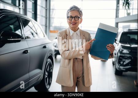 Femme professionnelle avec les cheveux gris courts pointe vers un dossier bleu tout en se tenant debout dans un concessionnaire automobile contemporain, entouré de véhicules élégants et Banque D'Images