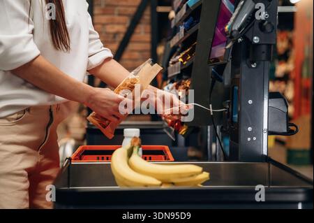 La cliente acheteuse à la caisse libre-service scanne les articles avec concentration, entourée de produits vibrants et de produits frais, illustrant la conve Banque D'Images