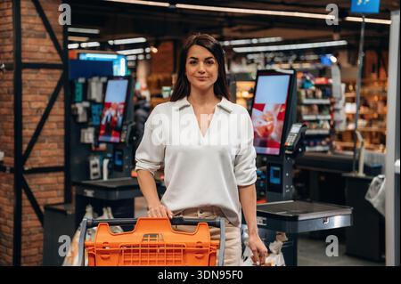Femme confiante avec des stands de cheveux foncés avec un panier dans une épicerie moderne, entourée de comptoirs de caisse et d'affichages de produits colorés, e. Banque D'Images