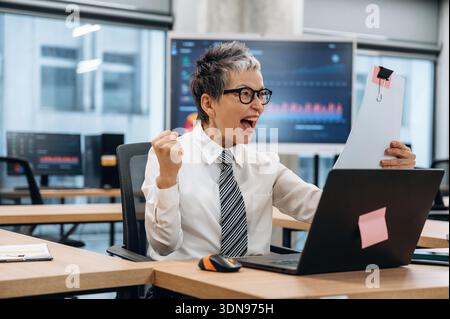 Femme d'affaires excitée dans un bureau moderne célèbre le succès tout en tenant un document, entouré par la technologie et un espace de travail dynamique, reflétant Banque D'Images