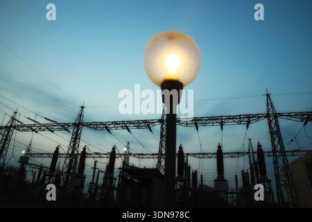 Un lampadaire lumineux se dresse au premier plan, silhouette contre un ciel crépusculaire. Derrière elle, une sous-station électrique complexe avec des tours et des fils i Banque D'Images