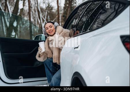 Jeune femme vêtue de vêtements chauds d'hiver souriant et tenant une tasse tout en étant assise dans une voiture avec une porte ouverte, entourée d'arbres enneigés, c Banque D'Images