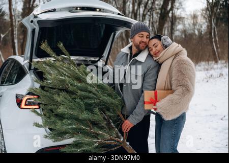 Femme asiatique et homme caucasien se préparent avec joie pour la saison des fêtes, chargeant un arbre de Noël et un cadeau dans le coffre de leur voiture, mis à nouveau Banque D'Images