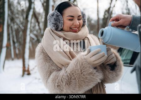 Femme heureuse portant des cache-oreilles et un manteau chaud tient une tasse tandis qu'une autre personne verse une boisson, dans un paysage enneigé avec des arbres, évoquant un sens Banque D'Images