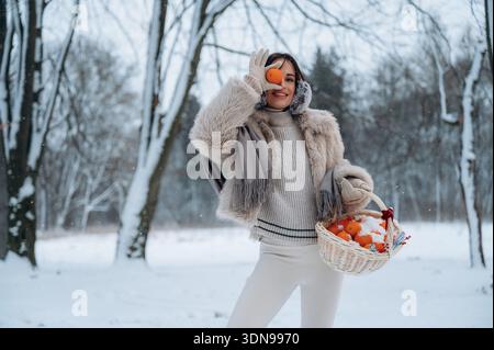 Figure féminine dans un manteau de fourrure tient une orange, debout dans un environnement enneigé avec des arbres, véhiculant un sentiment de joie et d'expérience de cueillette de fruits d'hiver Banque D'Images