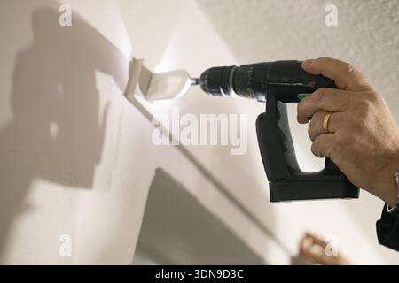 La main d’une personne utilisant une perceuse électrique sans fil pour installer un store au-dessus d’une fenêtre dans un espace intérieur. Rénovation domiciliaire. Banque D'Images