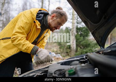 L'homme se tient près d'un capot de voiture ouvert dans une zone boisée. Il porte une veste de pluie jaune et verse du liquide dans le moteur de la voiture, se concentrant sur sa tâche pendant le dayli Banque D'Images