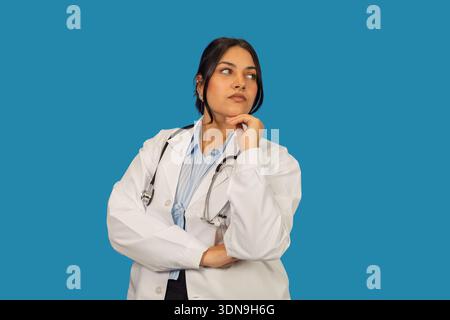 Professionnelle de santé féminine réfléchie portant une blouse de laboratoire et un stéthoscope sur un fond bleu Banque D'Images