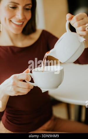 Une femme est assise à une table de cuisine et verse le thé d'une petite théière dans une tasse blanche. Elle sourit en profitant de sa pause de l'après-midi dans un espace confortable. Banque D'Images